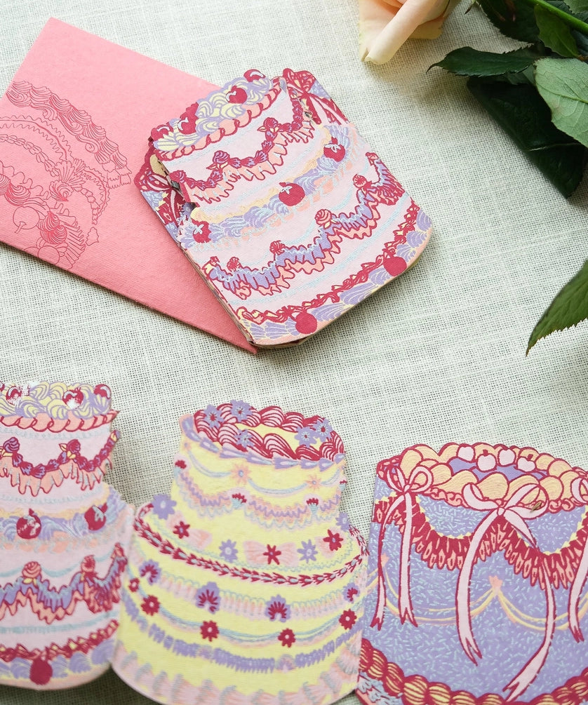 Still life image of East End Press Birthday Cake Concertina Card showing three of the four cake designs and the envelope. The cakes are decorated with various piping tips in colours like pink, purple, yellow, and red