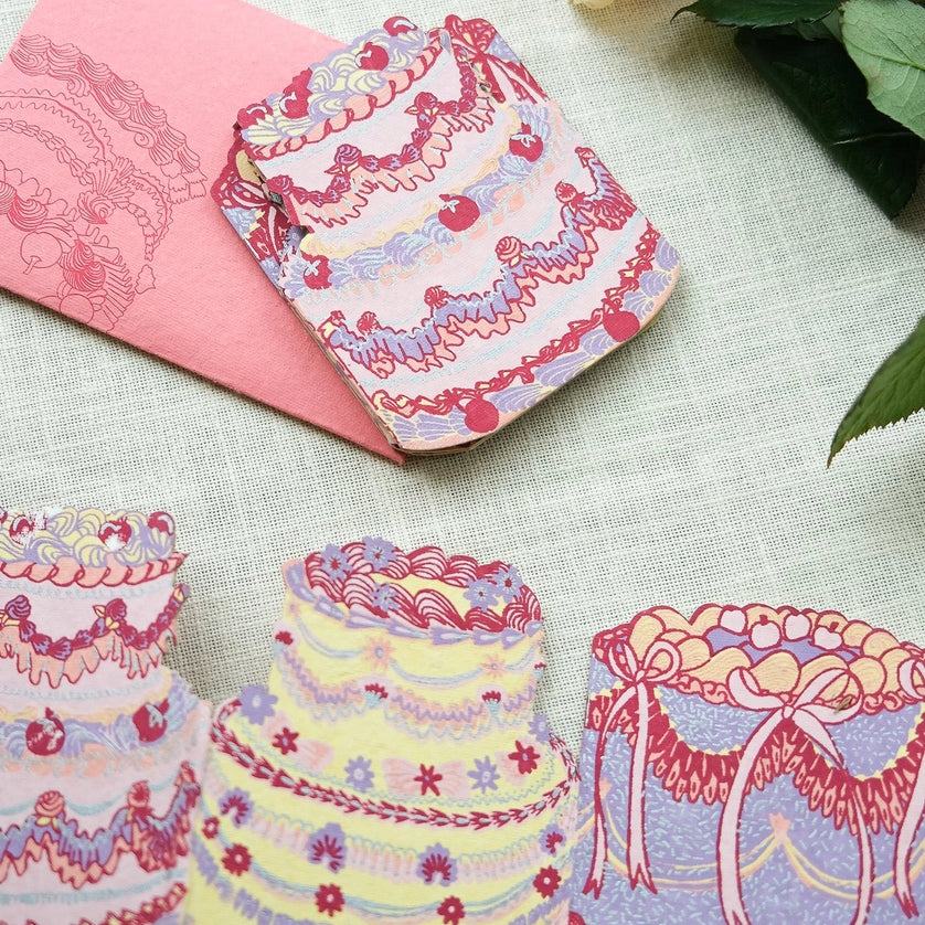 Still life image of East End Press Birthday Cake Concertina Card showing three of the four cake designs and the envelope. The cakes are decorated with various piping tips in colours like pink, purple, yellow, and red