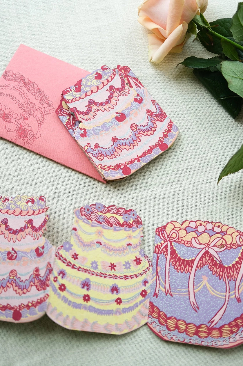 Still life image of East End Press Birthday Cake Concertina Card showing three of the four cake designs and the envelope. The cakes are decorated with various piping tips in colours like pink, purple, yellow, and red
