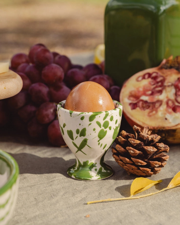 ceramic egg cup with a white base and moss toned green splatter design, photographed in a still life with an egg inside 