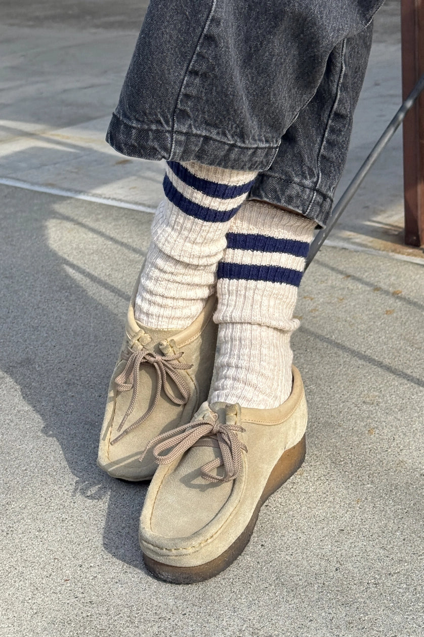 Person wearing beige shoes, Le Bon Shoppe Cottage Varsity Oatmeal socks, and dark jeans on a concrete surface.