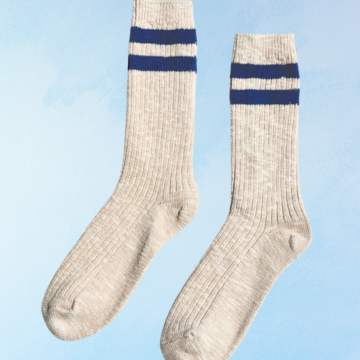Pair of off white socks with blue stripes on the top on a light blue background. Le Bon Shoppe Cottage Varsity socks in Oatmeal.
