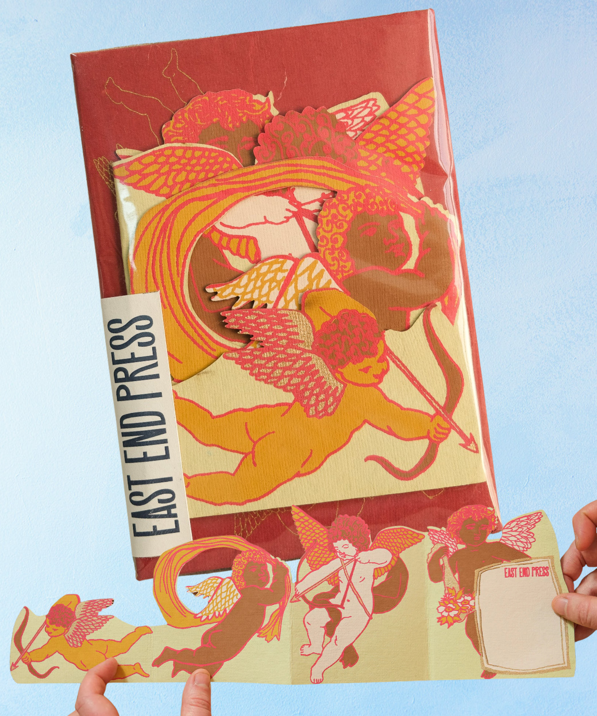 Packaged greeting card containing paper cherubs and 'East End Press' branding held against a blue background
