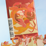 Packaged greeting card containing paper cherubs and 'East End Press' branding held against a blue background