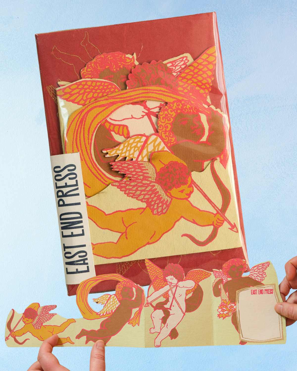 Packaged greeting card containing paper cherubs and 'East End Press' branding held against a blue background