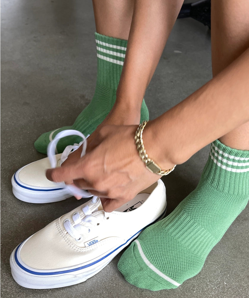 Person wearing green socks with white stripes and white sneakers on a concrete floor.