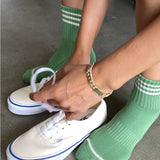 Person wearing green socks with white stripes and white sneakers on a concrete floor.