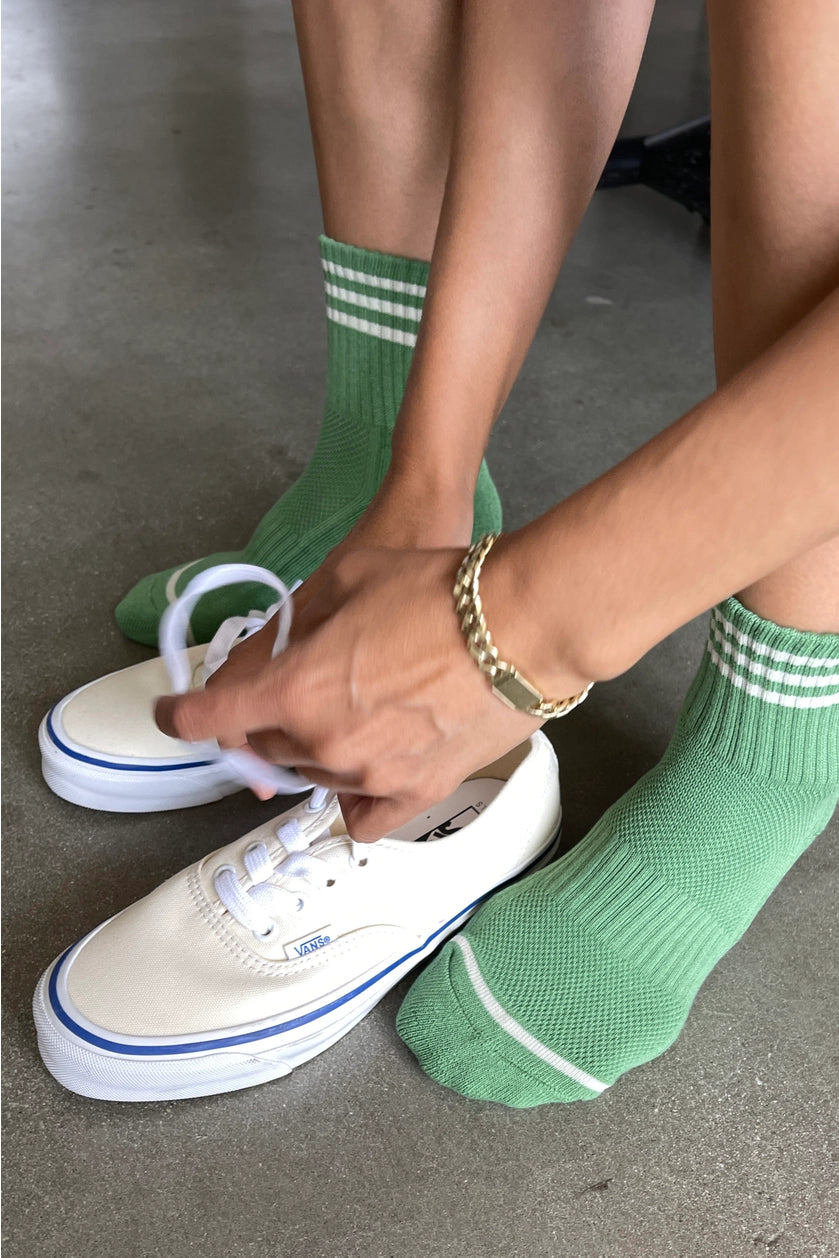 Person wearing green socks with white stripes and white sneakers on a concrete floor.