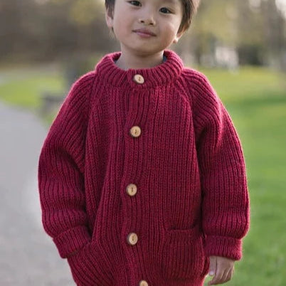 Little boy in a children's wool cardigan with a crew neck collar, patch pockets, english rib knit stitch body, and a finely detailed raglan sleeve.