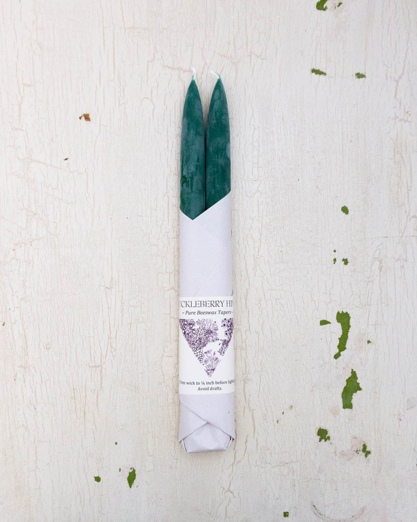 Set of two beeswax tapers in a dark green colour, bottom half wrapped in white paper by Huckleberry Hives.
