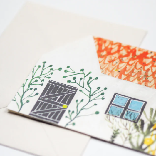A greeting card featuring perfect country cottage, with a cat in the yard and flowers around the front door. This is a house shaped card.