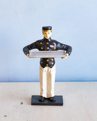 Male figurine of a bellhop on a small rectangular base,  in his arms he holds a large tray where cards can be placed. 