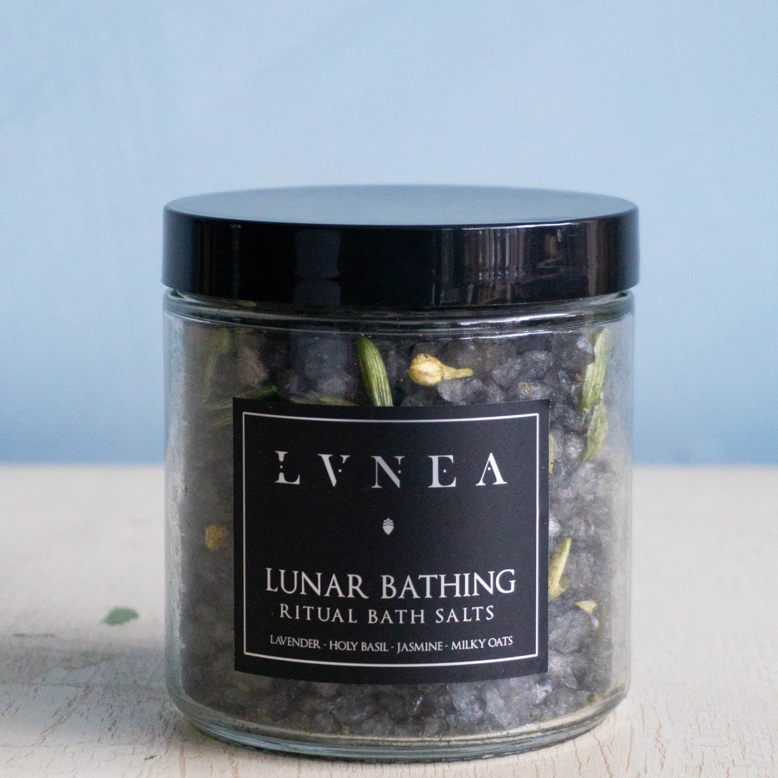 Clear glass jar filled with dark coloured coarse bath salts, dried lavender and jasmine flowers and dried oat tops by Lvnea.