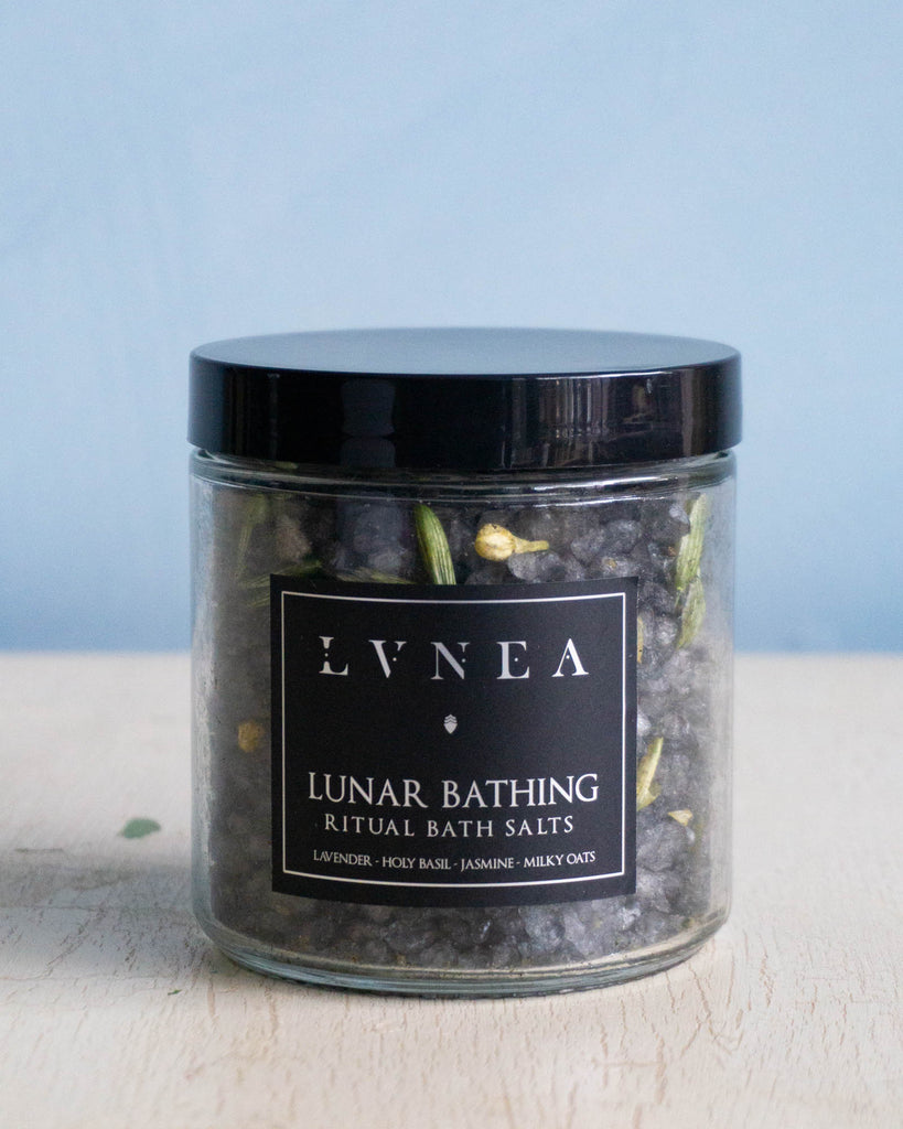 Clear glass jar filled with dark coloured coarse bath salts, dried lavender and jasmine flowers and dried oat tops by Lvnea.
