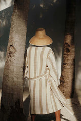 A woman standing backwards wearing a beige robe with yellow and brown vertical stripes, and a straw beach hat